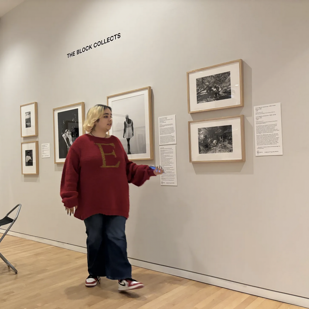 woman with dyed blond hair and red sweater with large E in center stands gesturing at photos on wall
