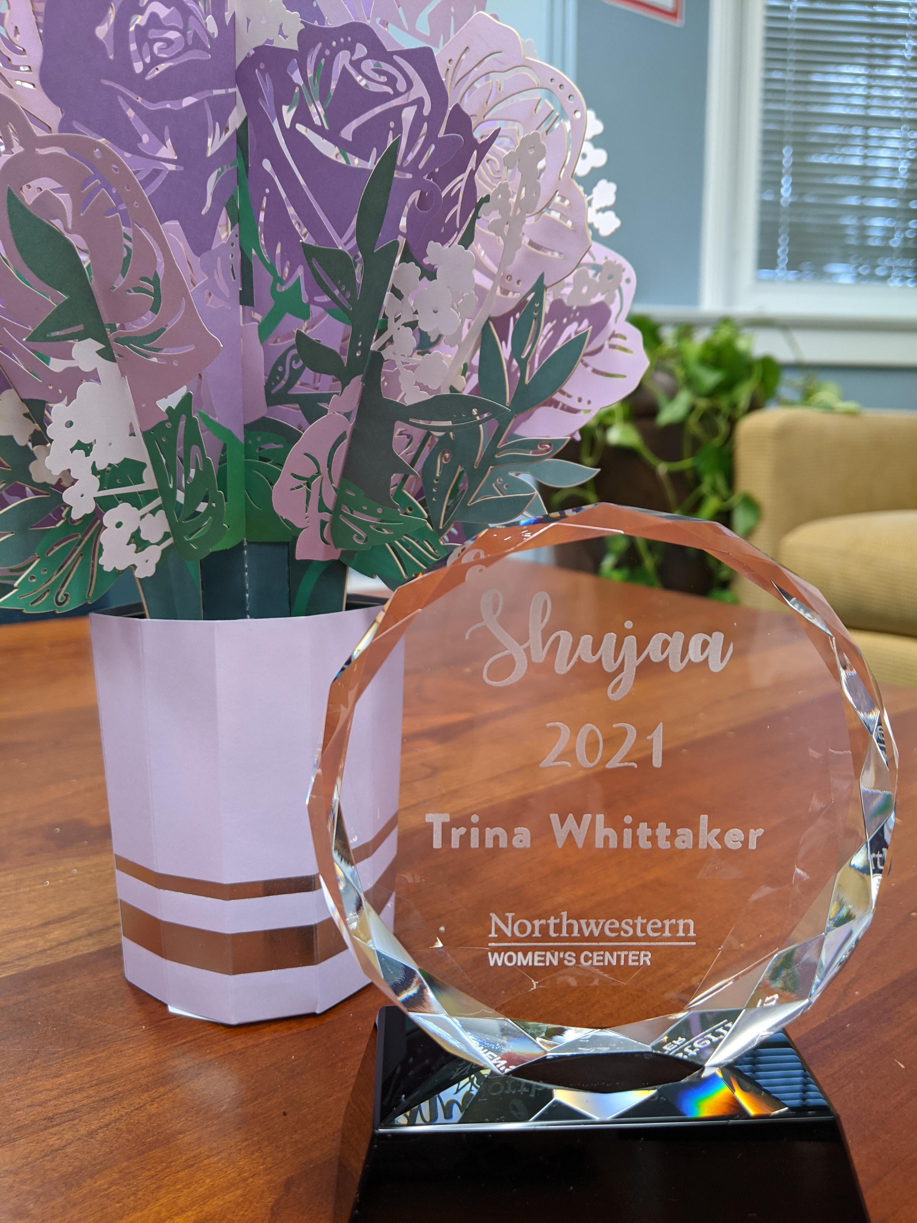 glass award on a table with paper flowers. Award texts is: shujaa 2021, Trina Whittaker. Northwestern Women's Center