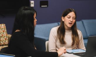 two students sitting at table, talking and looking at computer screen