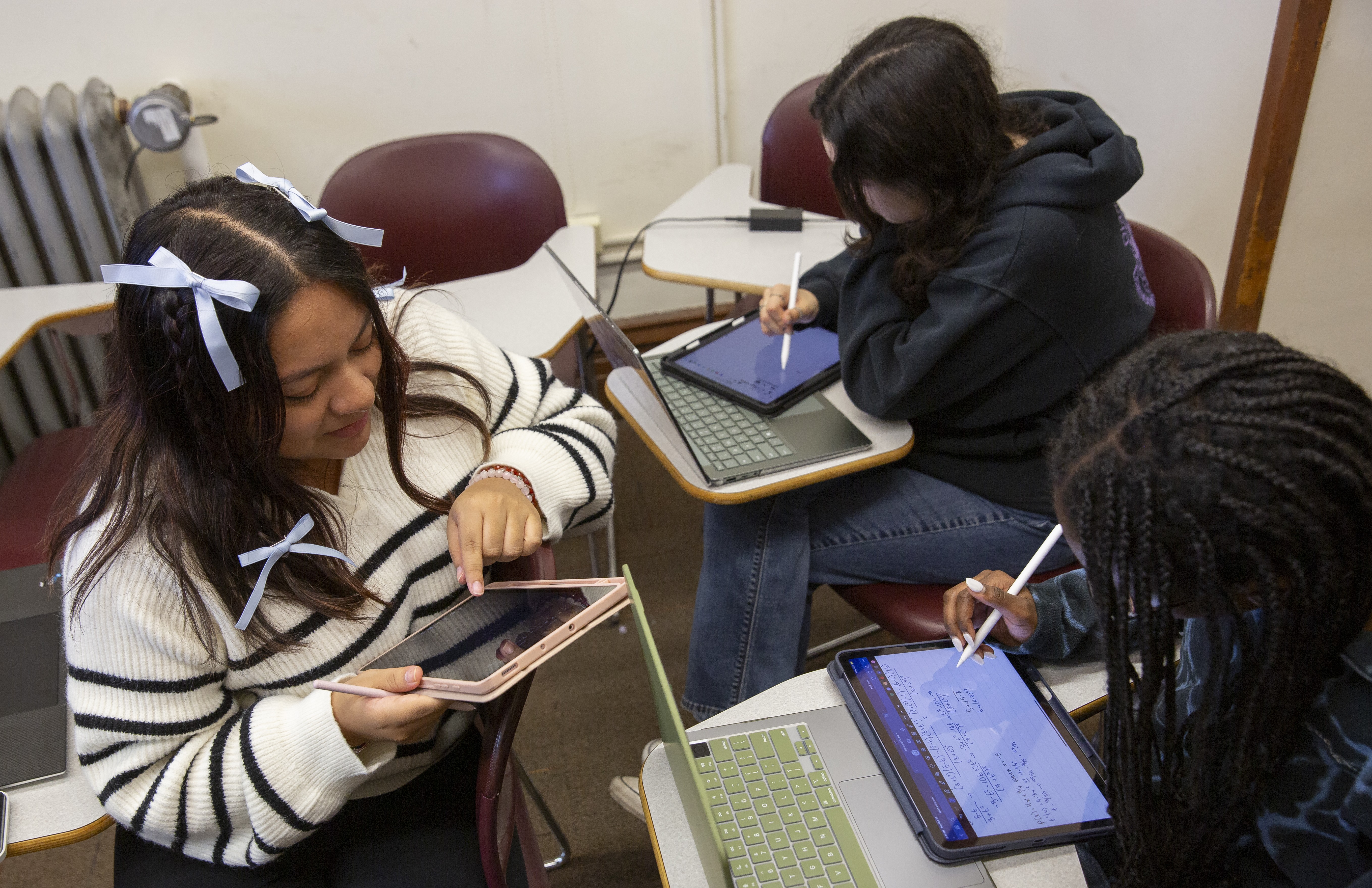 Three students in classroom, two seated and one kneeling, looking at computer tablet