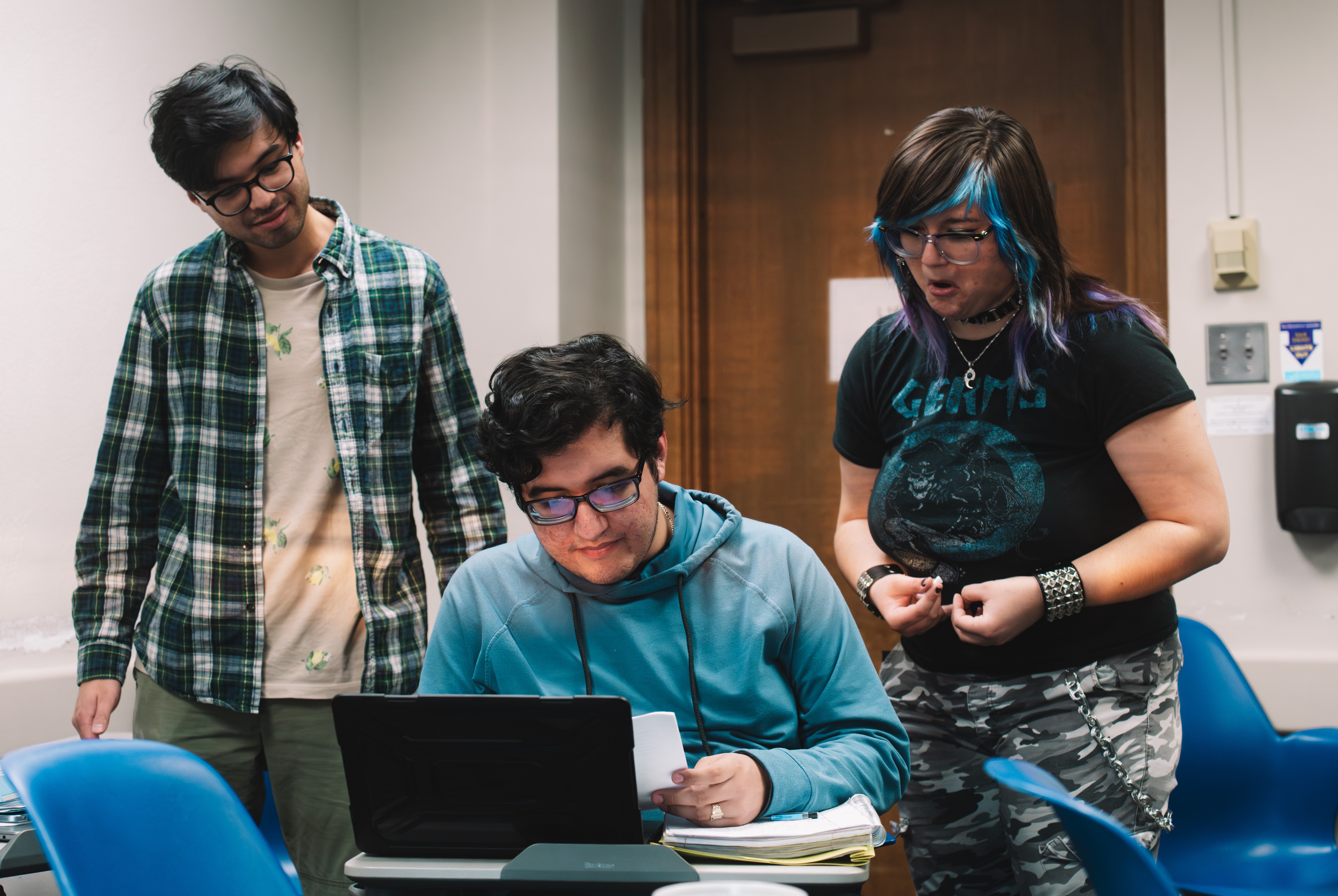 Three students in a classroom, two sitting and one standing, looking at a laptop