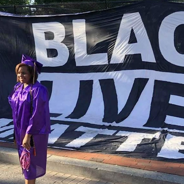 A black-identifying student stands in front of a Black Lives Matter banner in cap and gown.