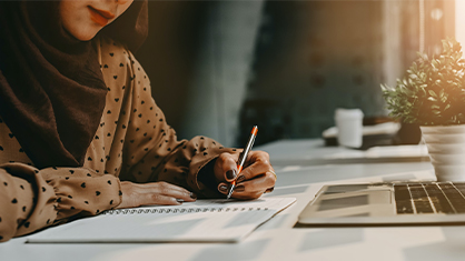 A person in a polka dot blouse writes in a notebook at a sunlit desk with a laptop and plant, conveying focus and productivity.