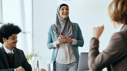 A woman in a hijab smiles while speaking to two colleagues during a workshop in a bright office. The mood is positive and collaborative.