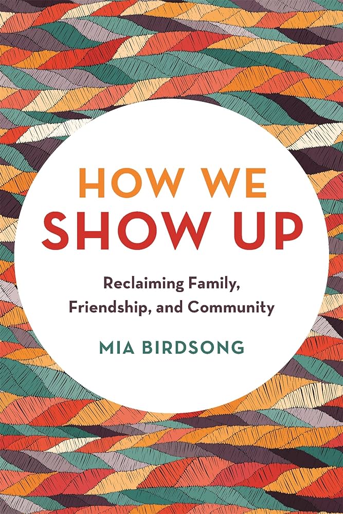 The cover of the book How We Show Up written in red and orange text that is placed inside a white circle. Beneath that text is multicolored braids interconnected with one another.