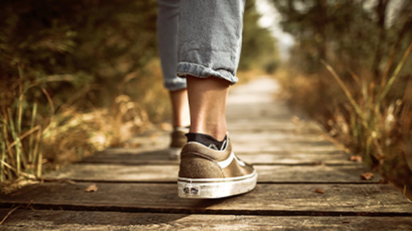 A low angle of person with blue jeans and brown  shoes on, walking alone on a wooden path.
