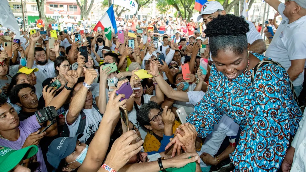 Francia Márquez, Colombia’s first Black Vice President, is shown standing on a stage wearing a traditional African print on a brown and blue dress, paying homage to her Afro-Latina heritage. Facing her is a crowd of people, seemingly excited, many of which are waving flags, others recording and taking pictures of her as she exits the stage.