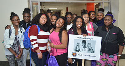 A group of young Black women pose together in a school hallway, smiling and holding a sign that reads “Wilma Rudolph – Black Female Summit” with a photo of the Olympic athlete. The image captures community, pride, and empowerment through representation and group identity.