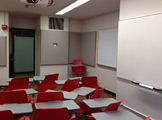 Picture of ooiking at the classroom and you can see a projector on the ceiling and two whiteboards on teh wall.