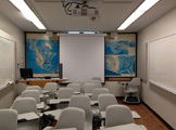 Picture of the front of the classroom with a projector and scree and maps on the wall