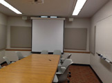 Picture of a long table with projector and screen and gray chairs