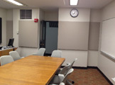 Picture of a long table with projector and screen and gray chairs with the teacher's podium