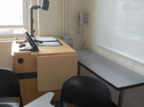 Photo of locy Hall Room Number 213 showing teachers desk, a lamp, phone on the wall and whiteboard