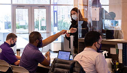 Students at a desk with several people sitting at the desk. They are being handed a card by a person working at the desk. Everyone is wearing medical facemasks.