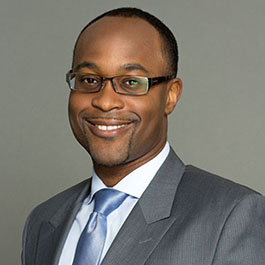 man with black hair and glasses in gray suit with blue tie smiling