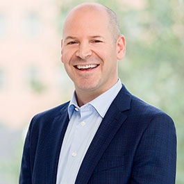 bald man in light blue shirt and dark blue jacket smiling