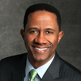 man with black hair blue shirt with green tie and gray suit smilig