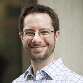 white man with dark hair and glasses wearing a patterned collared shirt and smiling