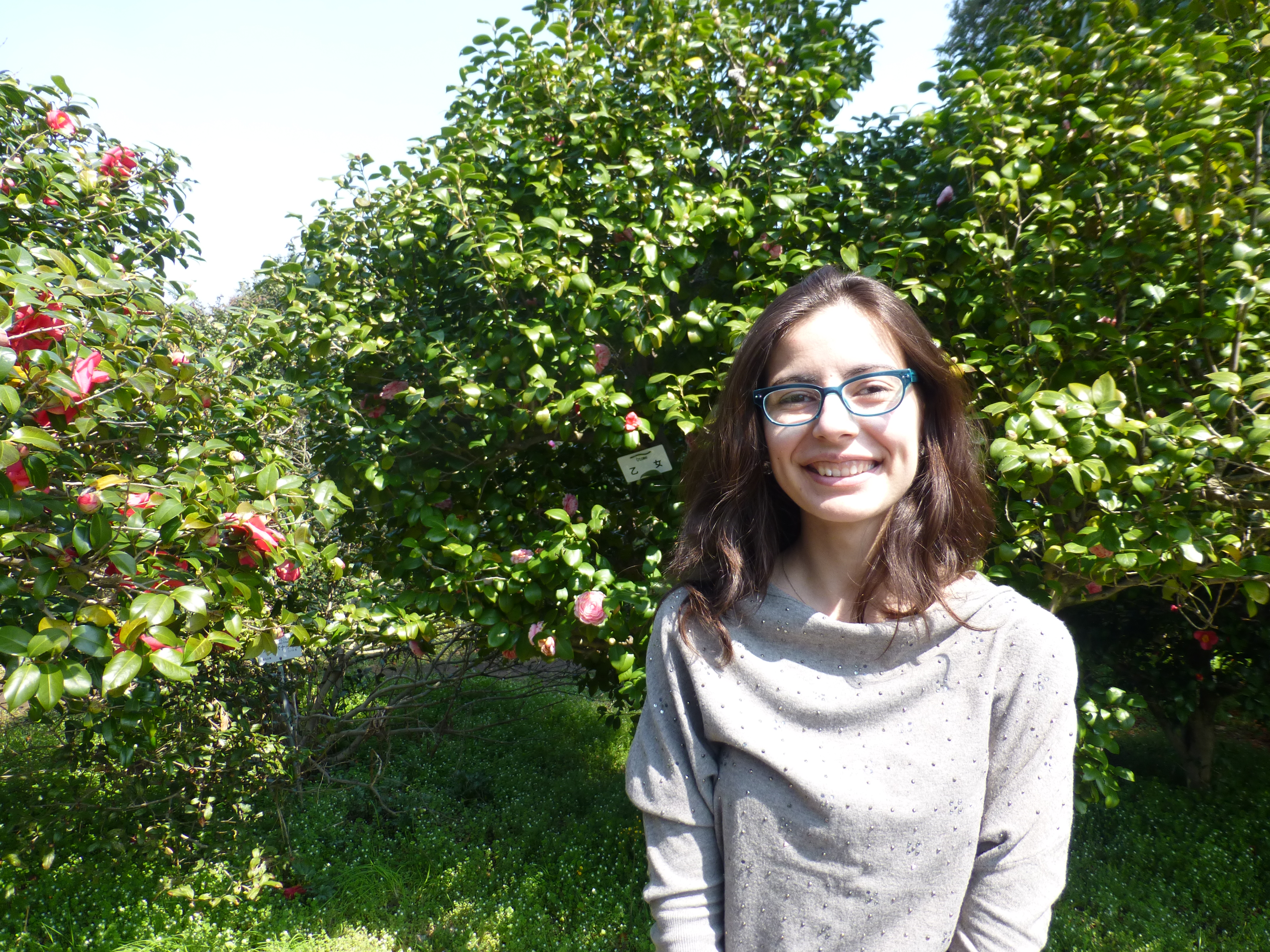 A woman smiles into the camera while standing in front of green trees.