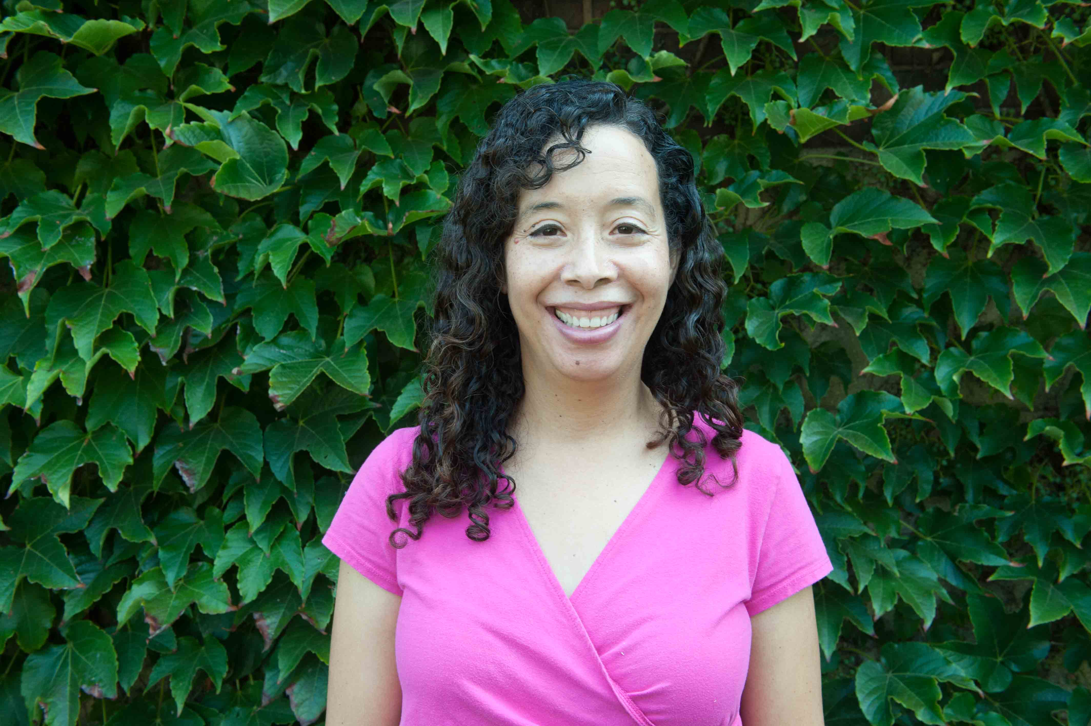 A woman smiles into the camera while standing in front of green ivy.