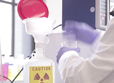 Lab worker wearing lab coat and gloves working near a waste container labeled Caution Radioactive Waste