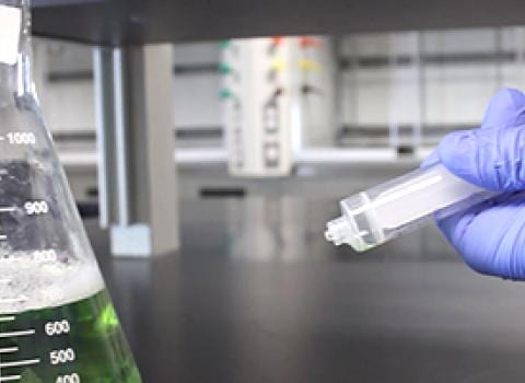 Gloved hand holding syringe near a beaker of green liquid on a lab bench