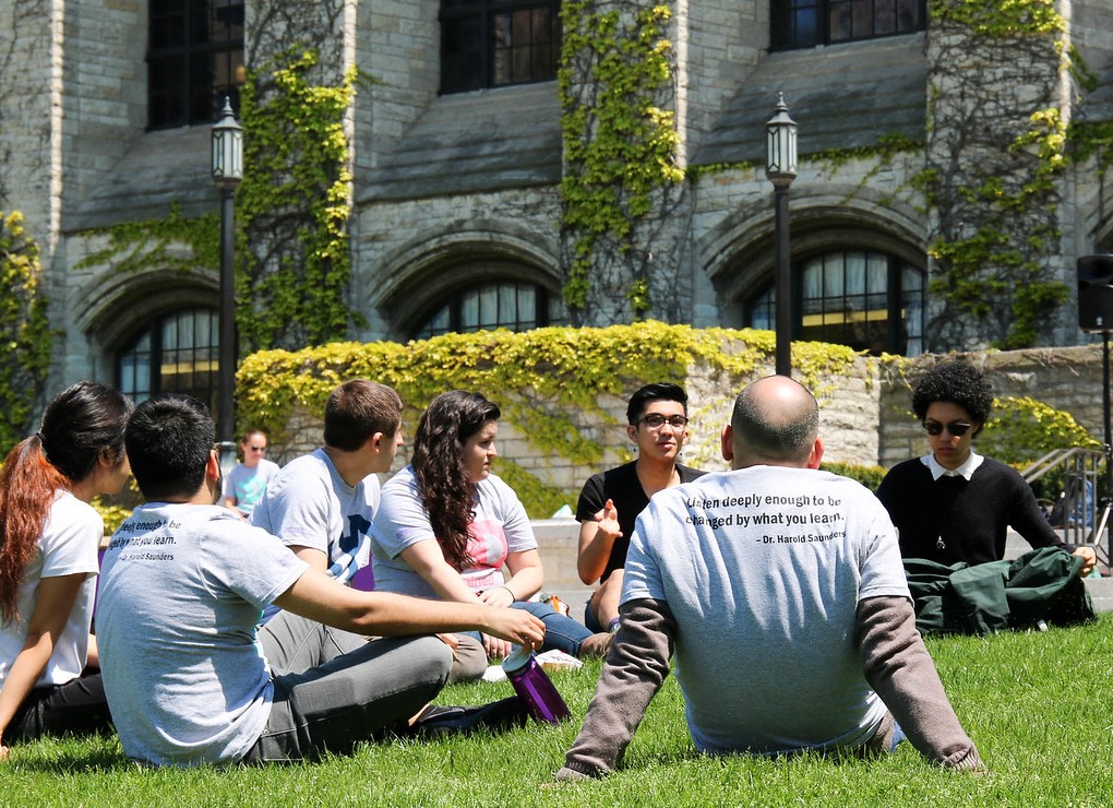 a group of students sit outside on a sunny day on the grass, having a discussion.