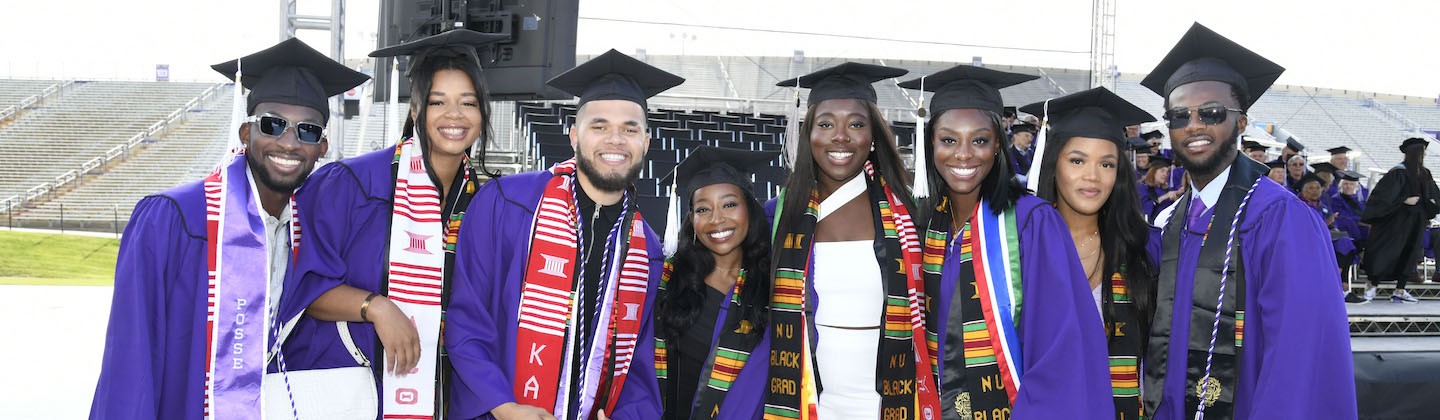 A group of students wearing purple regalia, black caps, and a variety of stoles stand together for a group photo