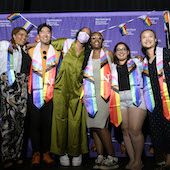 A group of students wearing stoles from Lavender Graduation pose for a photo in front of a purple banner that reads Northwestern Multicultural Student Affairs.