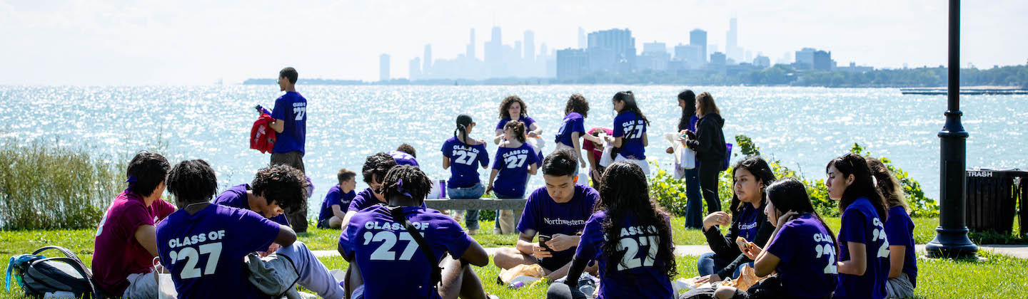 A group of students wearing purple shirts with "Class of '27" sit in circles eating lunch with a lake and Chicago city skyline in the background.