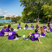 A group of students wearing purple shirts with "Class of '27" sit in circles eating lunch with a lake and Chicago city skyline in the background.