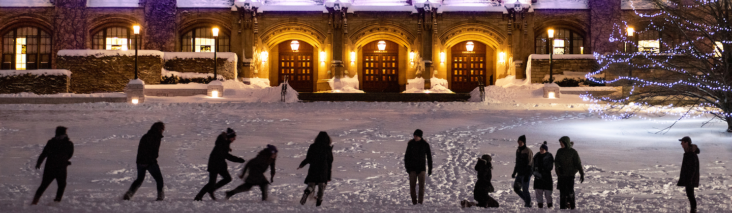 Students play in the snow at night with a building lit up in the background