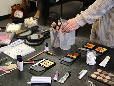 A collection of makeup is displayed as the instructor reaches for a brush.