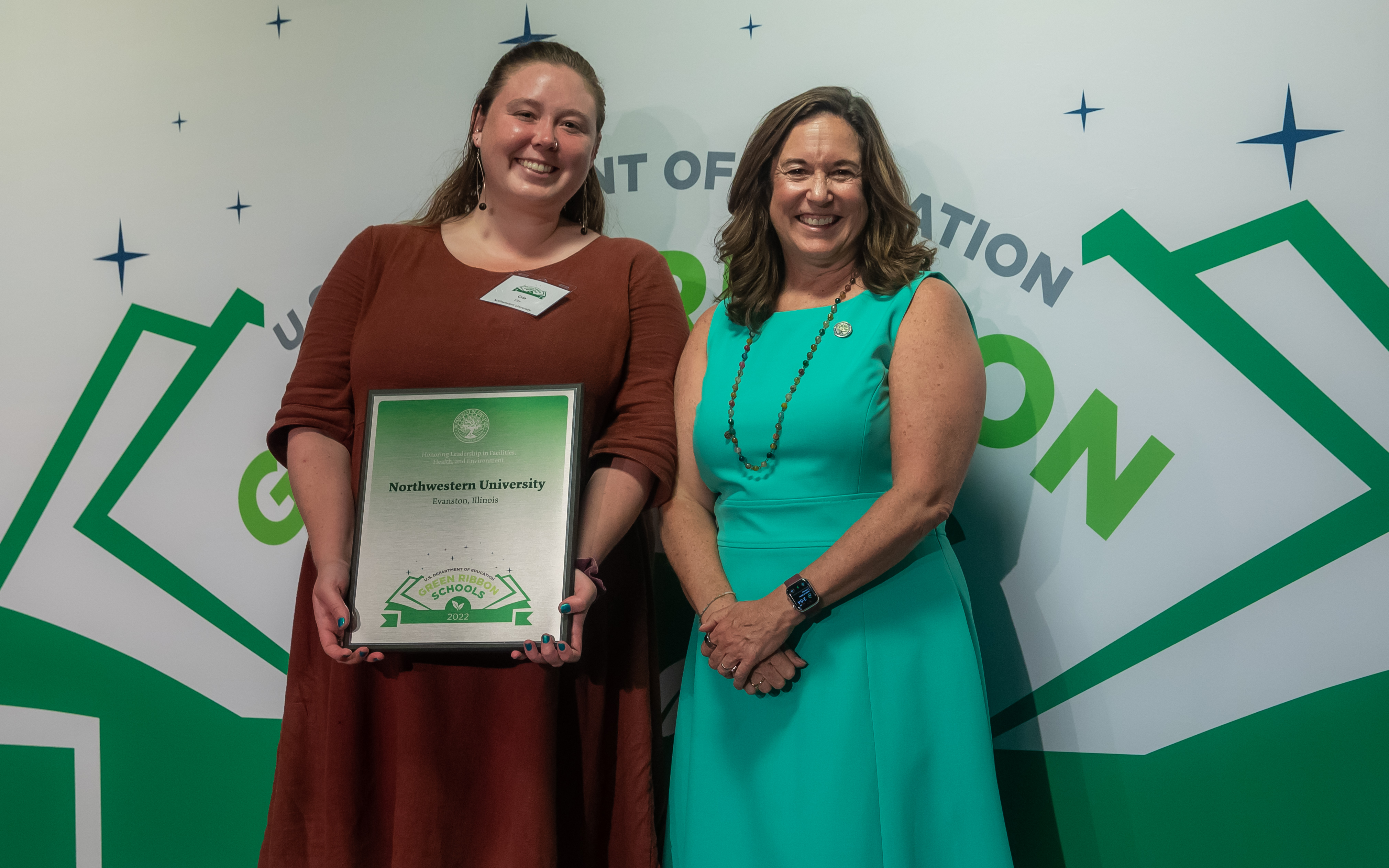 Two people, both with long hair and wearing dresses, hold an award at the 2022 Green Ribbon Schools Ceremony