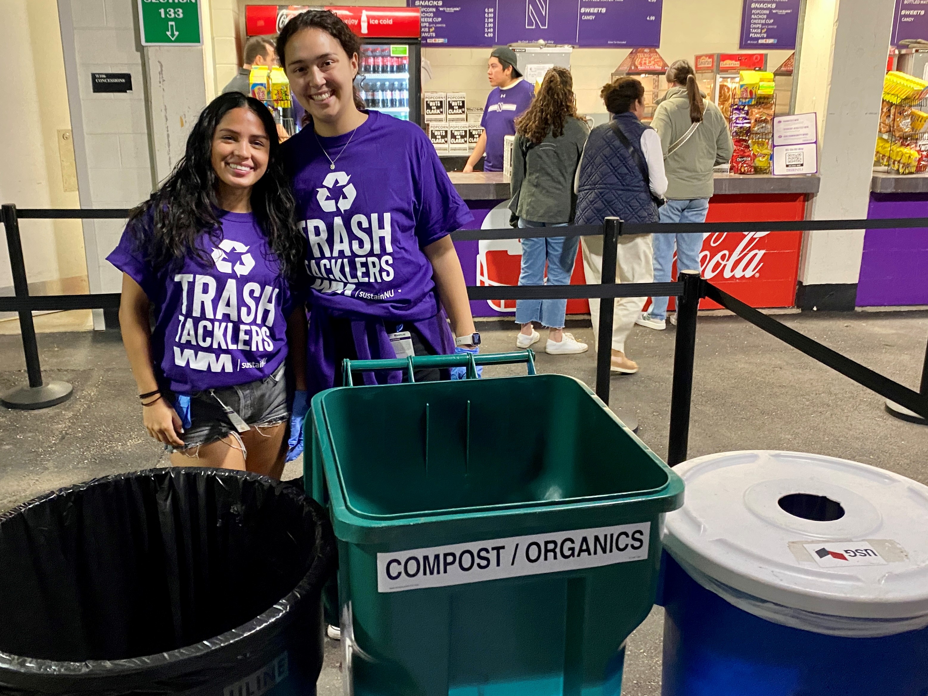 Two students in trash tacklers shirts stand over recycling, composting, and trash bins