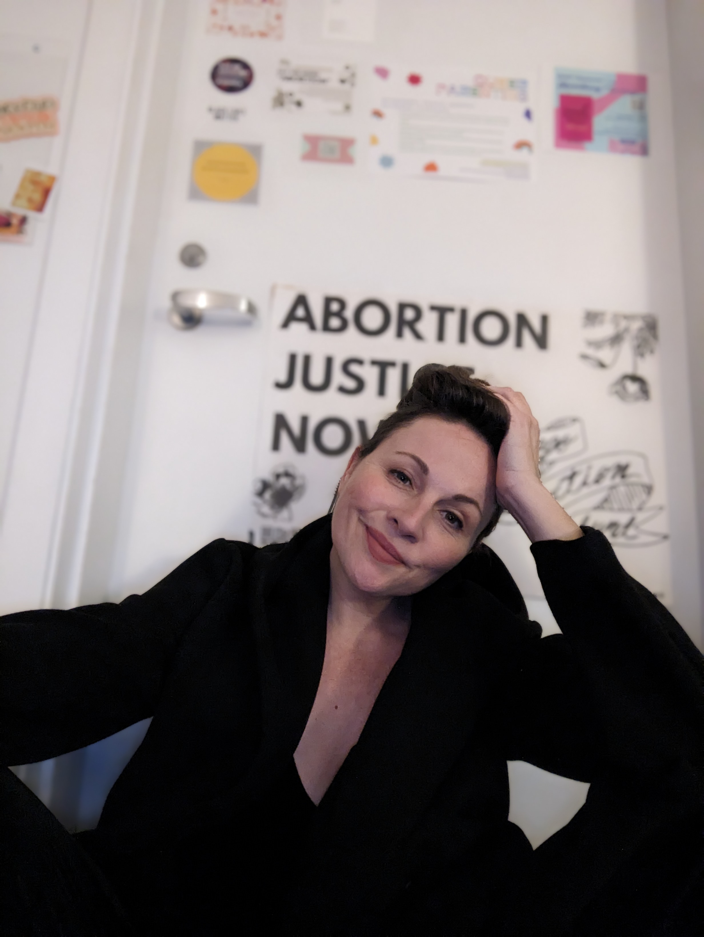 a white woman with brown hair and brown eyes sits in front of a door that reads abortion justice now. She is wearing a black coat and her hair is pulled back.