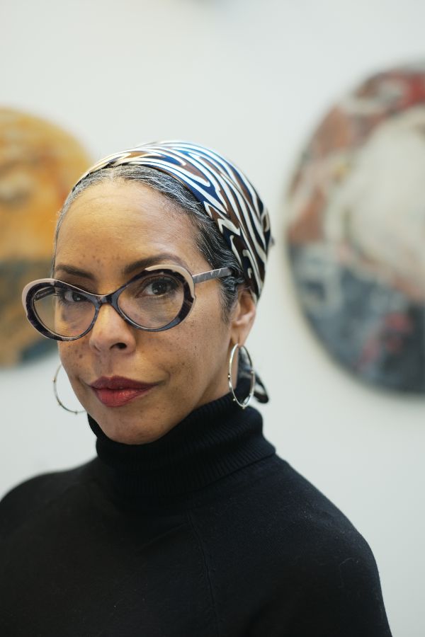 In the foreground is Kyrin, slightly turned left but looking into the camera. She is a Black women wearing a black turtle neck, silver hoop earrings, red lipstick, black framed glasses and a white, brown and blue scarf over her hair. In the background is blurred artwork hanging on a white wall. 